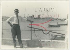 A man at a seafront area, while a ship and small boats are moored in the water